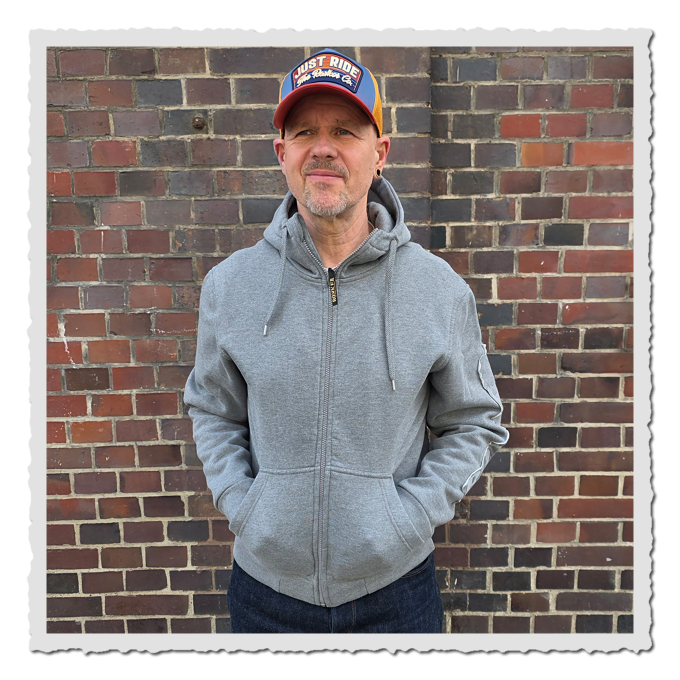 Person in hellgrauem Zip Hoodie von vorn vor Backsteinwand, mit Kappe, dunkler Jeans und Händen in den Taschen.